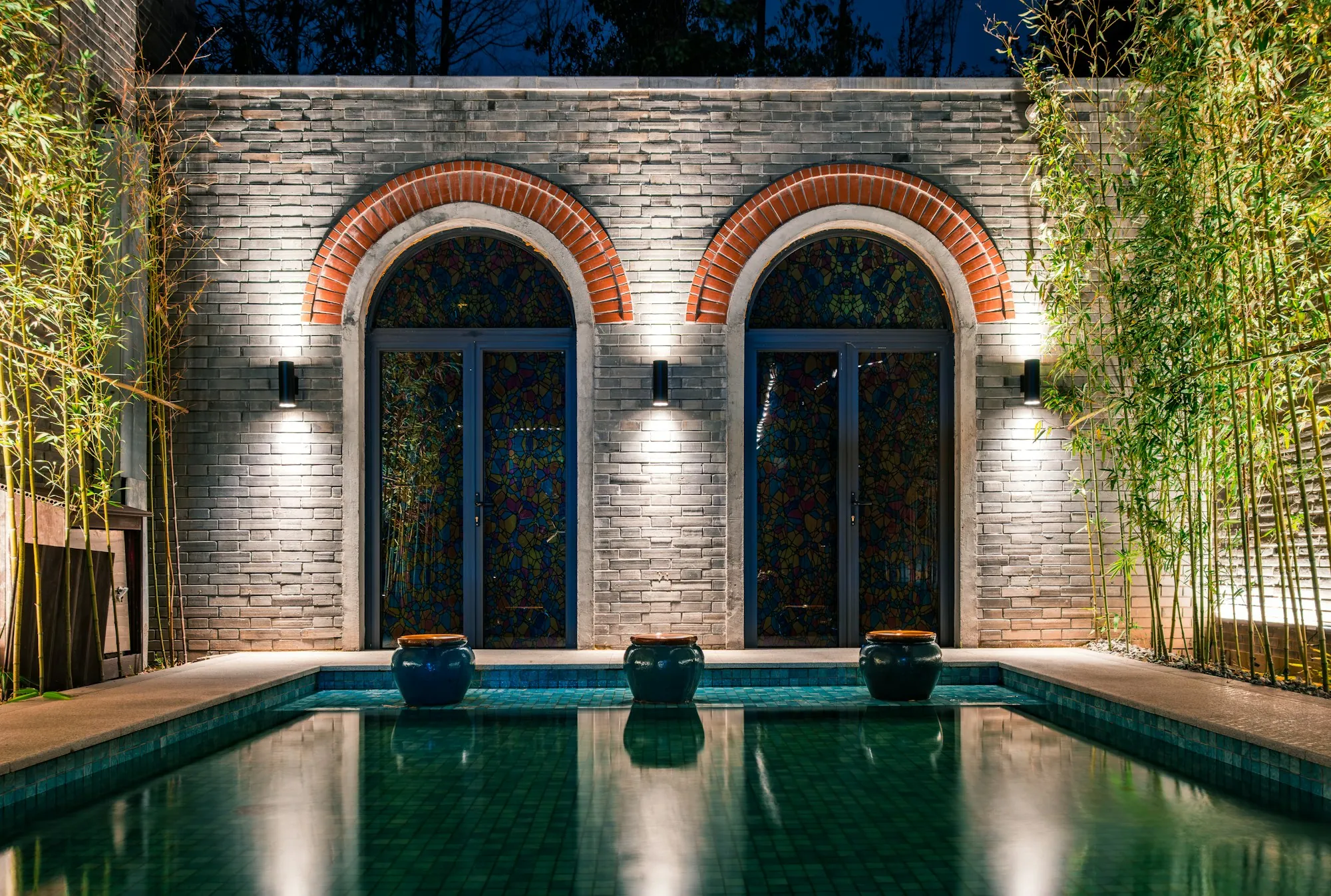 Piscine éclairée la nuit au pied de deux arches, entourée de bambous